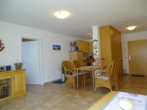 A dining area with wicker chairs and a table, a kitchen in the background, and a wooden cabinet with a lantern.