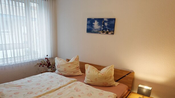 Quarto com cama, almofadas, mesa de cabeceira com abajur, pintura na parede e janela com cortinas.