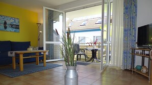 A living room with a sofa, a coffee table, a potted plant, a TV, and a sliding glass door leading to an outdoor area. - Comfortable vacation apartment in first beach row with balcony in Zingst - Residence (Zingst)