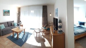 Un salon comprenant un canapé, une table à manger et des chaises, un téléviseur, et une chambre à coucher dans la pièce adjacente.