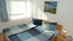 Un lit bien fait, avec une literie bleue et blanche, une tête de lit en bois et une table de chevet sur laquelle se trouvent une lampe et une petite décoration représentant une maison.
