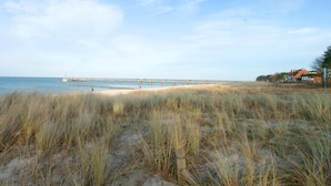 Ein Strand mit Dünen, einem Steg, der sich ins Wasser erstreckt, und ein Gebäude mit rotem Dach auf der rechten Seite.