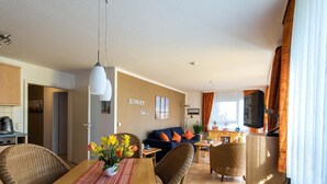 Een moderne woonkamer met een bank, een eettafel en stoelen. Er is een televisie aan de muur bevestigd en een raam met gordijnen.