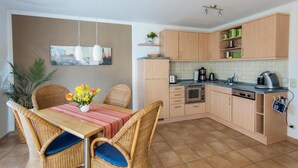 Een compacte keuken met houten kastjes, een eettafel met rieten stoelen en een klein aanrecht met een spoelbak.