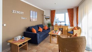 Een woonkamer met een bank, salontafel en rieten stoelen. Er is een keukenhoek met een aanrecht en kasten. De kamer heeft een beige muur met een bord waarop 'ZINGST' staat.