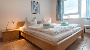 A neatly made bed with white linens and multiple pillows, a bedside lamp, and a small wooden nightstand with drawers. - Comfortable vacation apartment Residenz am Strand 1-11 in the first row to the Baltic Sea - (Zingst)