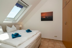 A bedroom with a skylight, a bed with white linens and blue towels, a nightstand with a lamp, a painting of a sunset, and a wooden wardrobe.