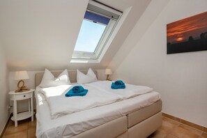 A bedroom with a skylight, a bed with white bedding, two blue towels, and a painting of a sunset on the wall. - Comfortable vacation apartment in the first row to the Baltic Sea with balcony in Zingst - (Zingst)