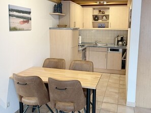 A compact kitchen with a dining area, featuring a wooden table and chairs, a small shelf with decorative items, and a tiled backsplash.