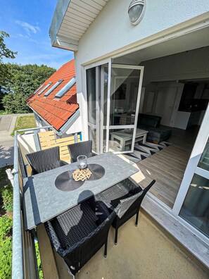 An outdoor dining area with a table and chairs, a glass door leading to a covered patio, and a red-tiled roof visible above. - Modern apartment in the first row on the beach of Zingst with balcony - Residence (Zingst)