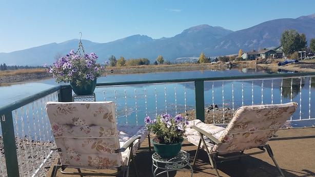Eine Terrasse mit zwei stuhlartigen Sitzgelegenheiten mit Blumenmuster und einem Blumentopf, mit Blick auf einen ruhigen See und ferne Berge.