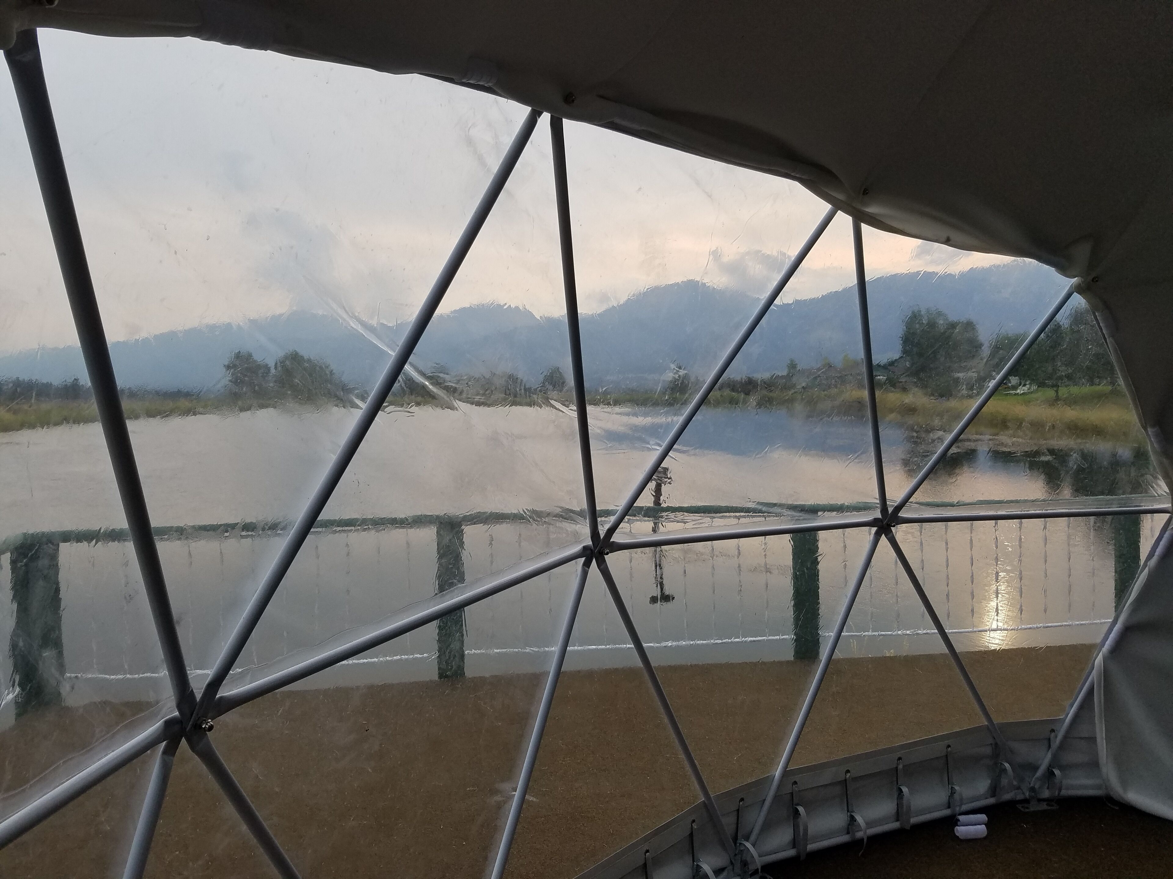 Vue de l’intérieur d’une structure ressemblant à une tente, dotée d’une armature métallique, donnant sur une étendue d’eau calme et des montagnes lointaines.