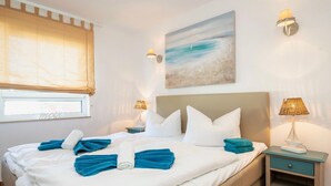 A neatly made bed with blue towels, a painting of a beach, and a nightstand with a lamp. - Wohnung 2-28 - Residenz am Strand (Zingst)