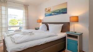 A neatly made bed with white linens and pillows, a painting of a boat on the sea above it, and two bedside lamps on matching nightstands. - Bright, comfortably furnished apartment directly on the dyke and beach - Residence (Zingst)