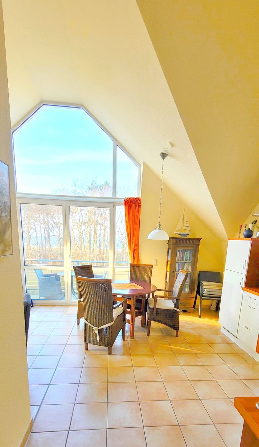 A spacious room with a high ceiling, large windows, a dining area with chairs, a small table, and a kitchen area with cabinets.