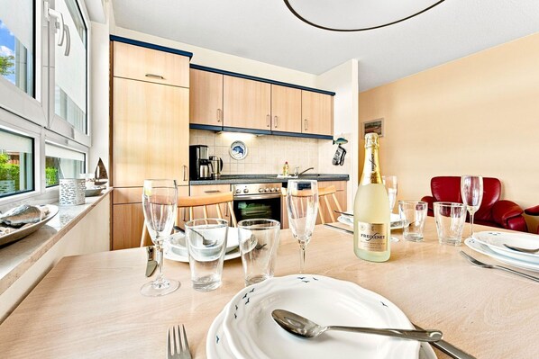 A modern kitchen with wooden cabinets, a dining table set for six with wine glasses and a bottle of champagne, and a red leather chair in the background.