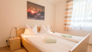 A double bed with white bedding, a nightstand with a lamp, and a painting of a sunset on the wall.
