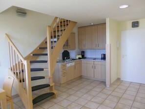 Una cocina con armarios de madera, lavavajillas y estufa. Se observa una escalera de madera con escalones negros que conducen a un nivel superior.