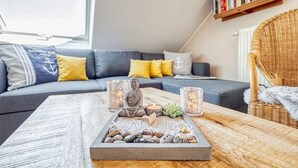 Una sala de estar acogedora con un sofá gris, cojines amarillos, una silla de mimbre y una bandeja con velas y una estatua de Buda.