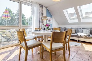 Ein Essbereich mit rundem Tisch, vier Stühlen und einer Couch mit Kissen. Zwei Fenster mit Vorhängen, ein Balkon mit buntem Sonnenschirm und ein Oberlicht über der Couch.