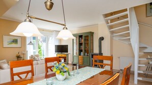 Ein Essbereich mit Holztisch, Stühlen und einer Blumenvase. Eine Treppe befindet sich rechts, im Hintergrund ist ein Fernseher zu sehen.