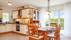 Eine moderne Küche mit weißen Schränken, ein Essbereich mit Holzsesseln und große Fenster mit Vorhängen.
