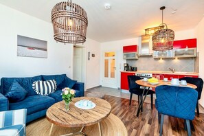 Un salon moderne avec un canapé bleu, une table basse en bois et un coin repas, avec une cuisine rouge en arrière-plan.