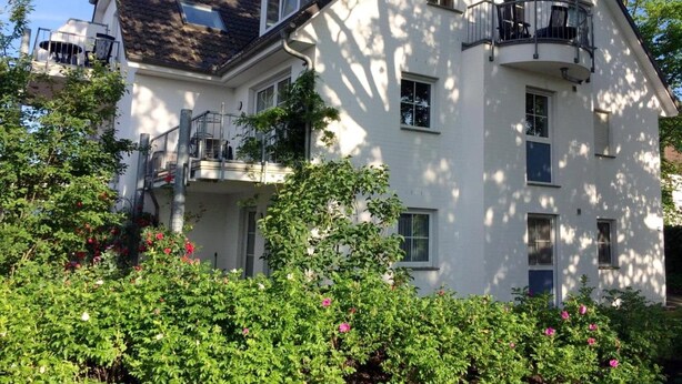 Une maison à deux étages dotée d’un balcon, entourée d’une végétation luxuriante et de plantes en fleurs.