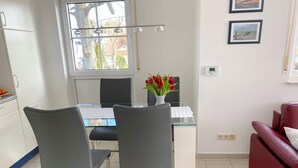 Une cuisine moderne avec des meubles blancs, une table à manger avec des chaises, un canapé rouge et une fenêtre donnant sur des arbres et des immeubles.