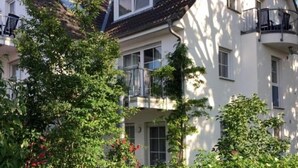 Une maison à deux étages avec un balcon au deuxième étage, entourée d’une végétation luxuriante et de plantes en fleurs.