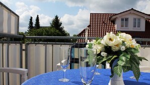 Une table avec deux verres à vin vides et un vase de fleurs blanches, le tout posé sur une nappe bleue, avec en arrière-plan une maison à toit de tuiles et une clôture.