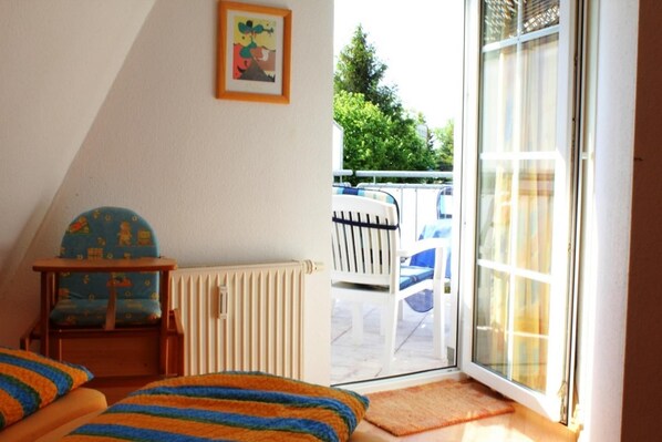 A room with a sloped ceiling, a radiator, a small table with a chair, a colorful rug, and a sliding glass door leading to an outdoor area with a chair and a table.
