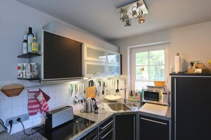 Een moderne keuken met een inbouwoven, magnetron en spoelbak. Er zijn kastjes, een raam en een aanrecht met diverse keukenbenodigdheden.