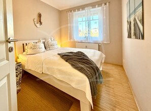 Une chambre à coucher comprenant un lit, des oreillers, une couverture, un radiateur, une fenêtre avec des rideaux et un élément de décoration fixé au mur.
