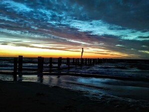 Ein Strand mit einer hölzernen Mole, die sich ins Meer erstreckt, Wellen, die an das Ufer schlagen, und einem Sonnenuntergang im Hintergrund.