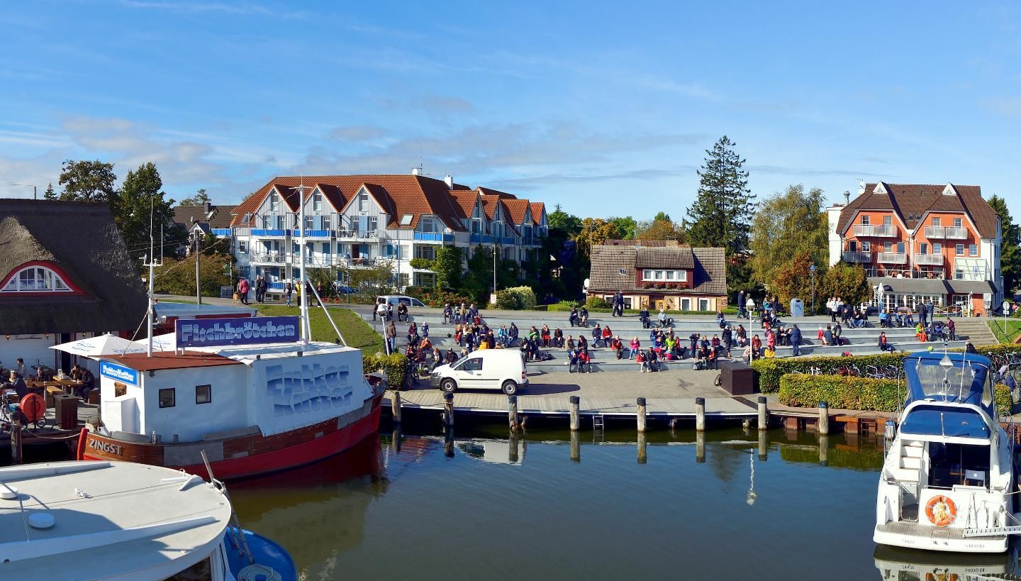 Uma marina com barcos atracados, um cais com pessoas e edifícios ao fundo.