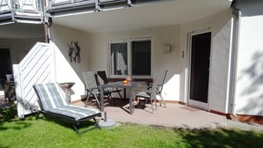 Un patio con una mesa y sillas, una silla lounge a rayas y un pequeño jardín.