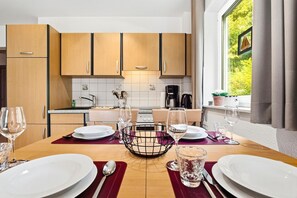 A modern kitchen with wooden cabinets, a dining table set for two with wine glasses, and a view of greenery through the window. - Sobek Vacation Apartment - Bright first-floor apartment with a furnished, covered terrace (Zingst)