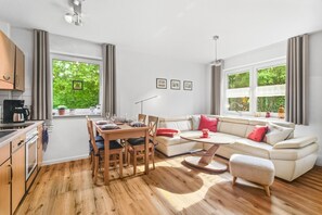 A modern living room with a dining area, large windows with curtains, and a view of greenery.