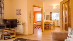 Una sala de estar con televisor, radiador, un macramé colgado en la pared, y una puerta que da a un comedor con mesa y sillas.