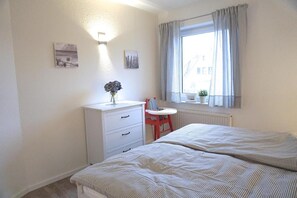 Ett sovrum med två sängar, ett litet skrivbord, en stol, ett element och ett fönster med gardiner.