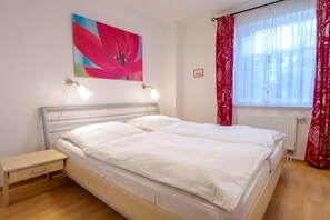 A double bed with white linens, a bedside table, a wall-mounted lamp, a window with red patterned curtains, and a floral painting on the wall. - Comfortable vacation apartment with terrace and garden in a central location in Zingst - (Zingst)