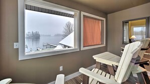 Een kamer met een groot raam met uitzicht op een meer, een tafel met stoelen, en een aanlegsteiger en bomen in het zicht.