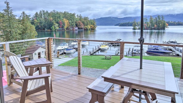 Een houten terras met een tafel en stoelen, met uitzicht op een jachthaven vol boten en een meer, omringd door bomen en bergen.