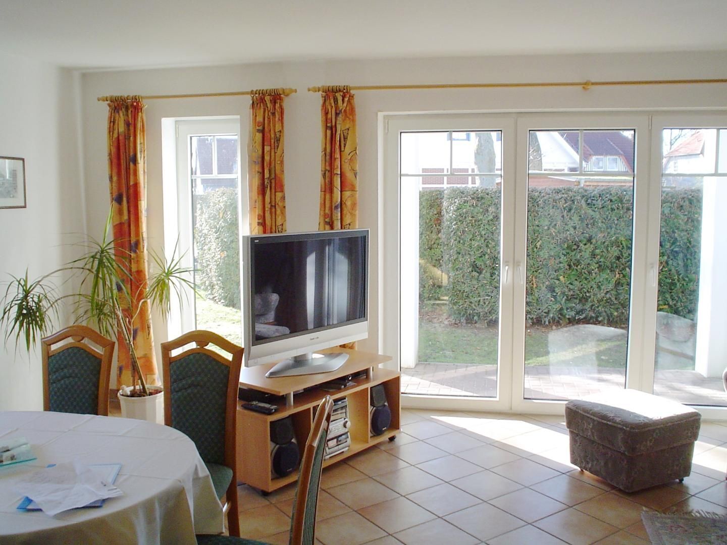 A living room with a television on a wooden stand, a dining table with papers, two chairs, a potted plant, and a sliding glass door leading to a garden.