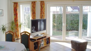 A living room with a television on a wooden stand, a dining table with papers, two chairs, a potted plant, and a sliding glass door leading to a garden.