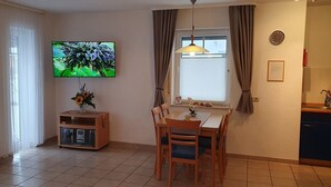 A dining area with a TV mounted on the wall, a table with chairs, a vase with flowers, and a window with curtains.