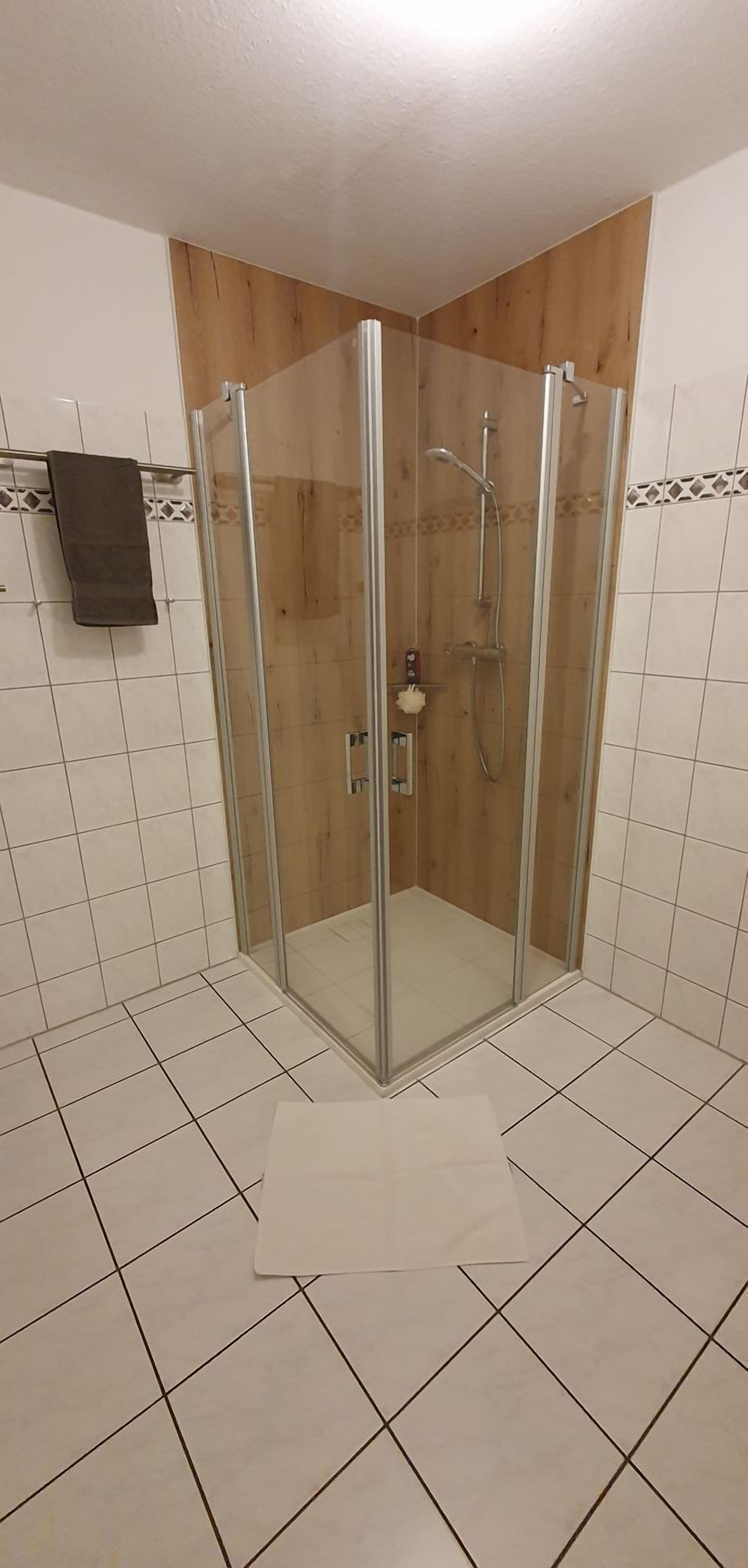 A shower area with a glass door, tiled floor, and wooden paneling on the walls.
