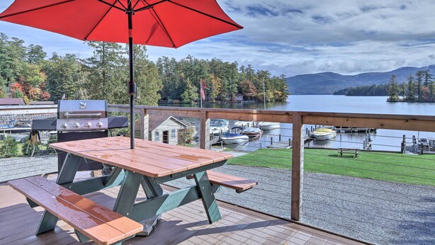 Une terrasse en bois avec une table de pique-nique et des bancs, donnant sur un lac où l’on peut voir des bateaux et un quai.