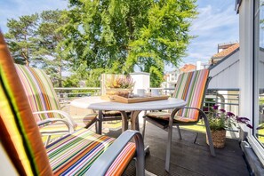Eine Terrasse mit gestreiften Stühlen und einem Tisch, umgeben von einer Brüstung und Blick auf Bäume und Gebäude.